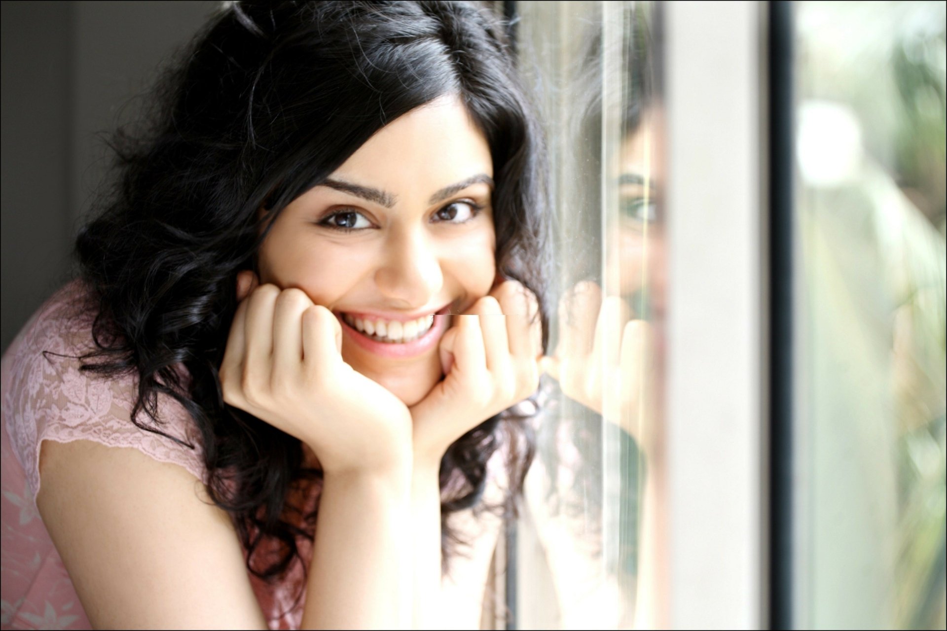 A vibrant HD wallpaper featuring celebrity Adah Sharma, smiling warmly with her hands resting on her chin, captured by a window, radiating happiness and charm.