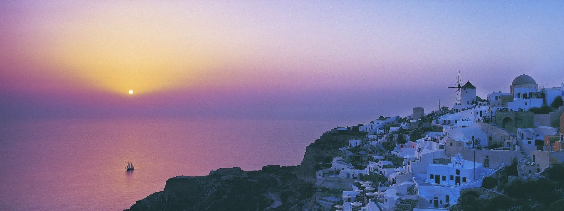 HD desktop wallpaper of Santorini's iconic white buildings on a cliff at sunset, overlooking the calm sea under a soft pastel sky.