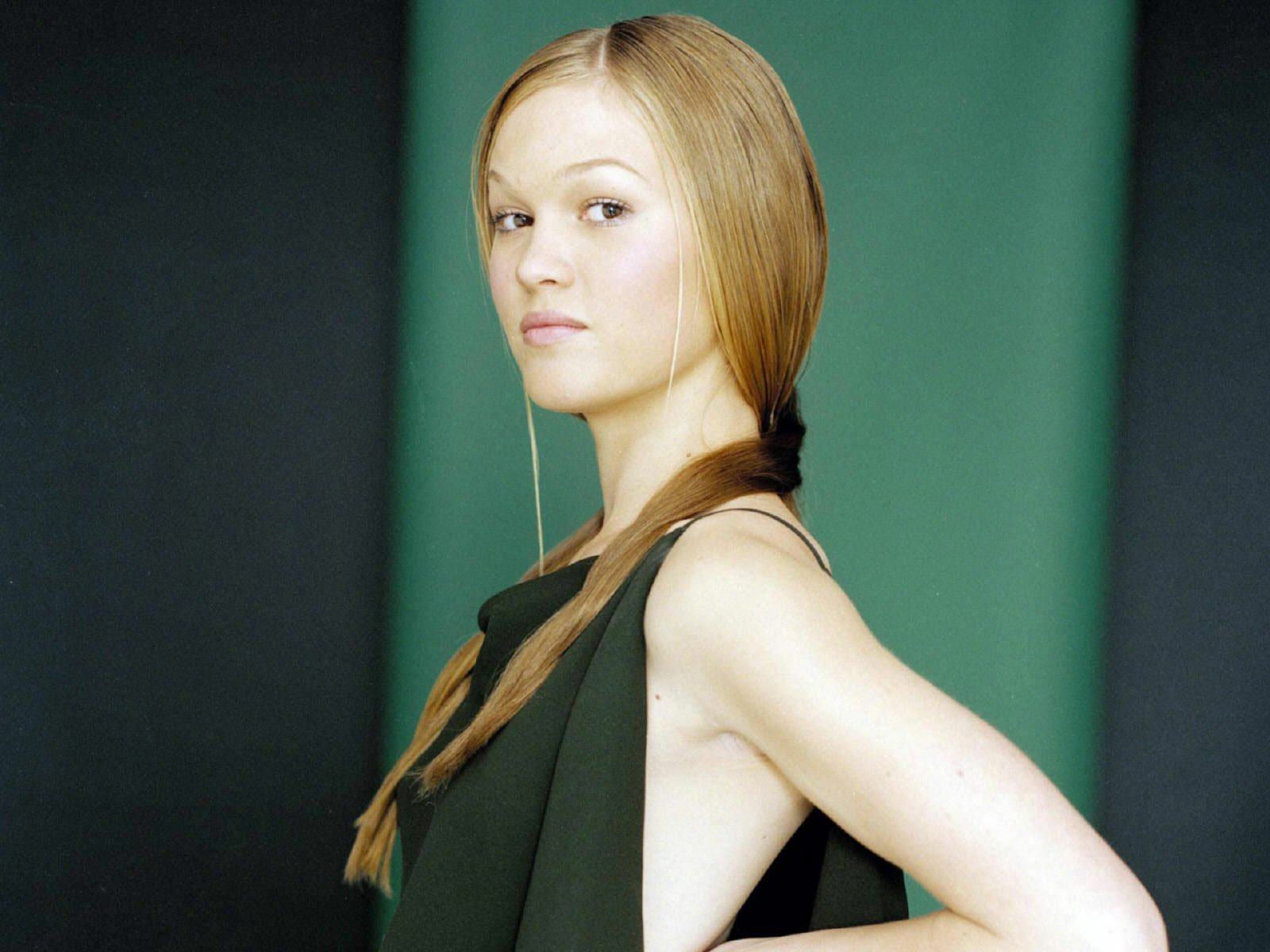 A striking HD desktop wallpaper of Julia Stiles, featuring her with long, straight hair styled in a relaxed braid, confidently posing against a muted green background.