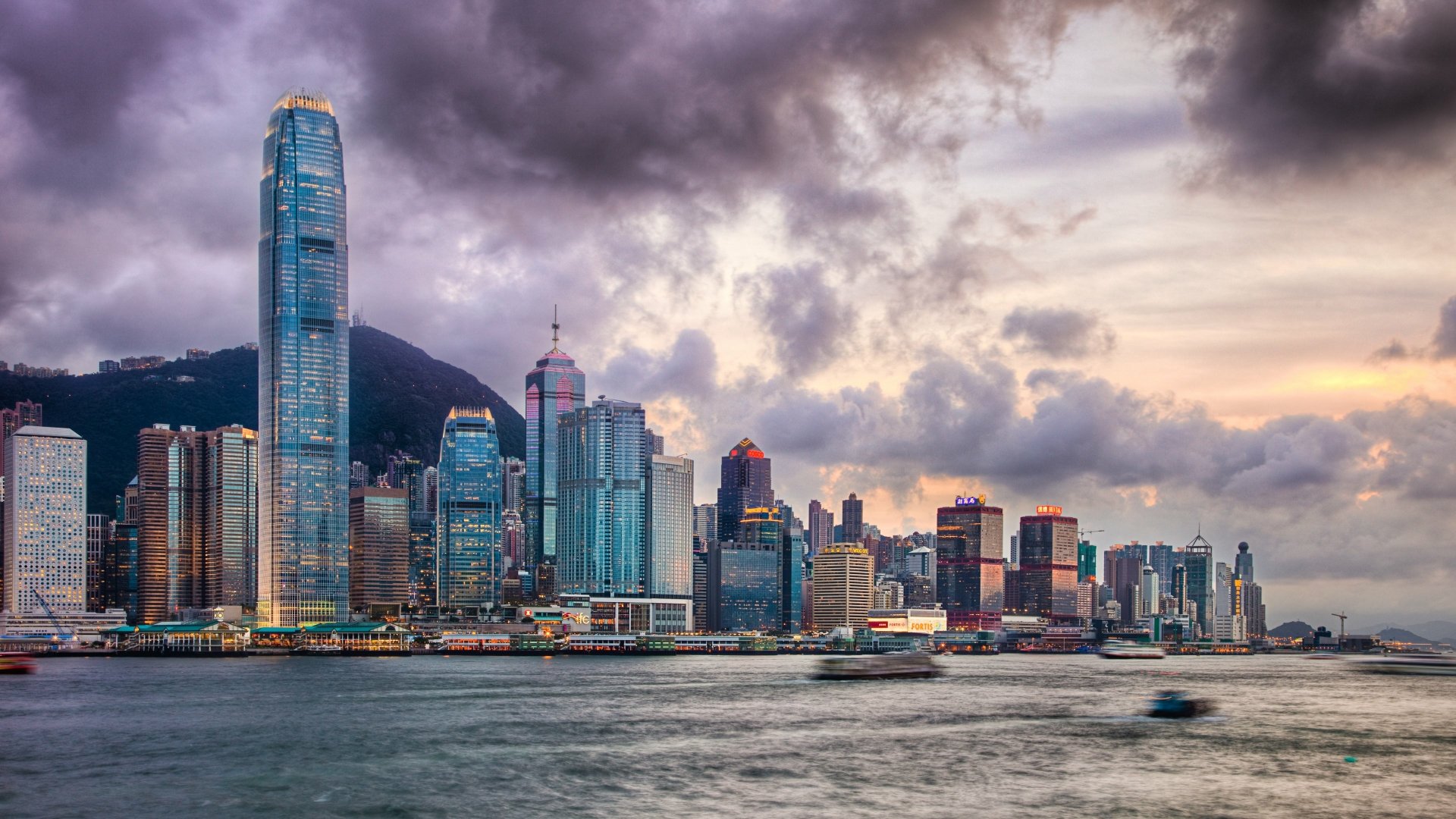 4K Ultra HD wallpaper of Hong Kong’s man-made skyline at dusk, featuring towering skyscrapers under dramatic cloudy skies along the waterfront.