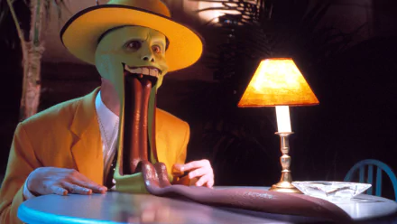 HD desktop wallpaper featuring The Mask character with an exaggerated grin, portrayed by Jim Carrey, in a yellow suit and hat, sitting at a table with a lamp.