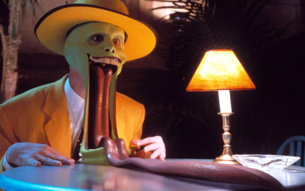 HD desktop wallpaper featuring The Mask character with an exaggerated grin, portrayed by Jim Carrey, in a yellow suit and hat, sitting at a table with a lamp.