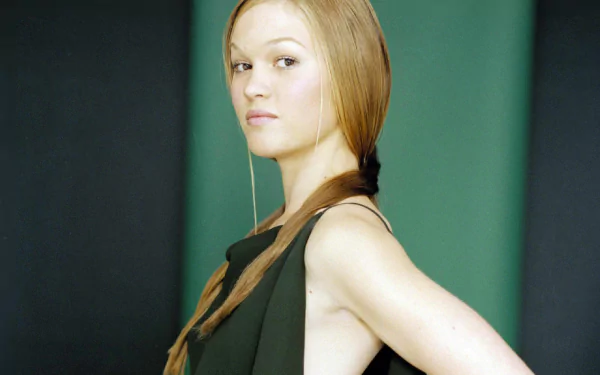 A striking HD desktop wallpaper of Julia Stiles, featuring her with long, straight hair styled in a relaxed braid, confidently posing against a muted green background.