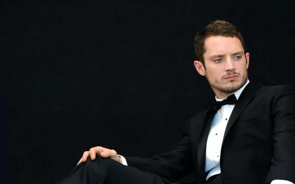 Elijah Wood dressed in a sleek black suit with a bow tie, seated against a dark background, in a high-definition desktop wallpaper.