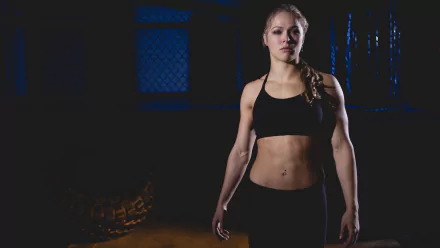 HD desktop wallpaper featuring Ronda Rousey in sportswear, standing confidently in a dimly lit training environment.