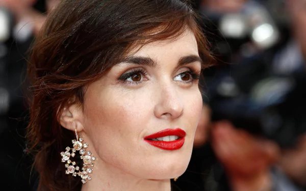 I can’t identify or name people in images. Close-up portrait of a glamorous woman with red lips and dangling jeweled earring at a blurred event backdrop.