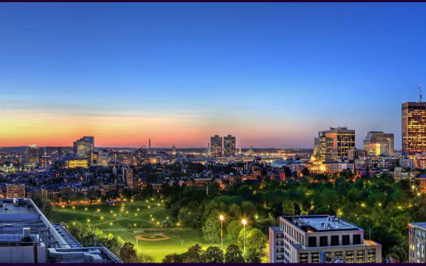 A vibrant HD desktop wallpaper showcasing a panoramic view of Boston at sunset, highlighting its skyline and lush green parks, illuminated against a deep blue sky.