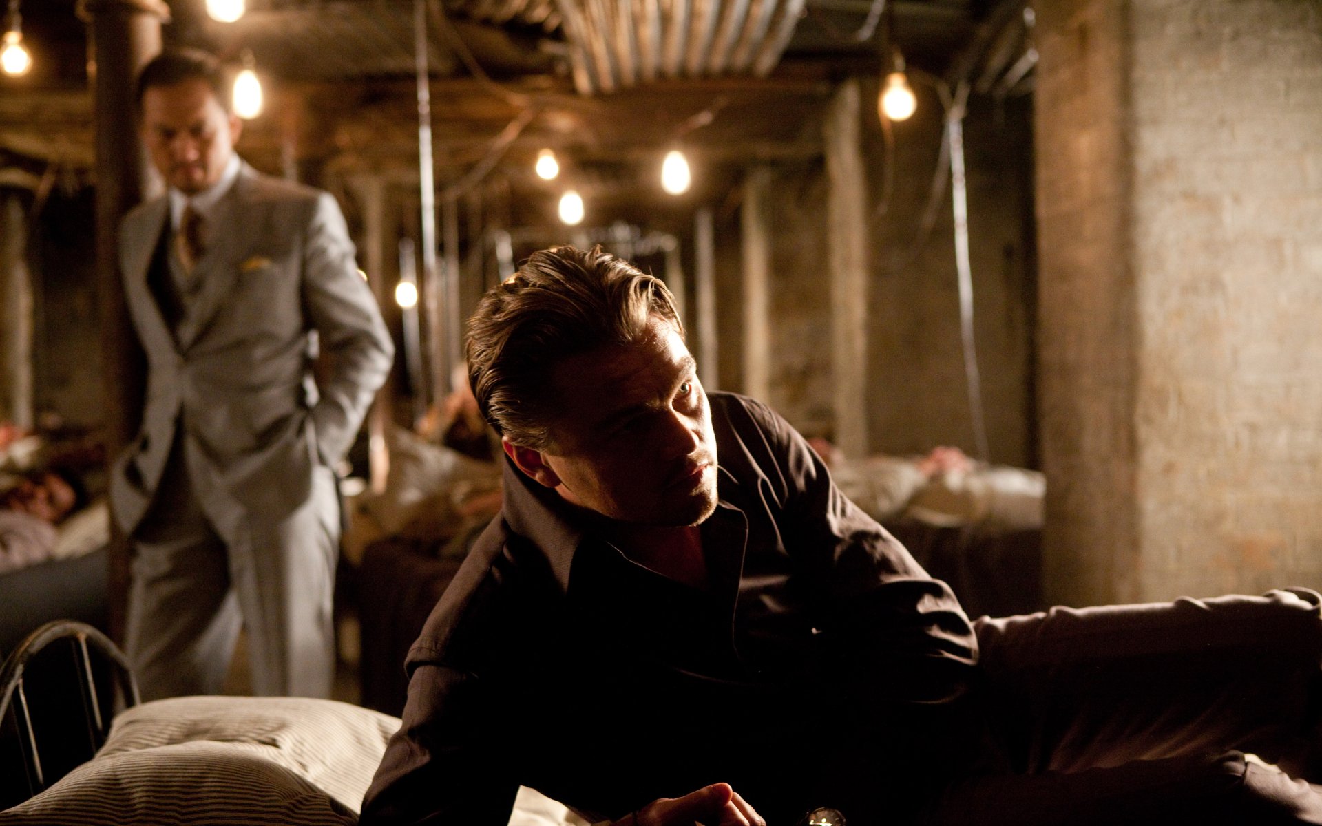 4K Ultra HD PC desktop wallpaper: Cobb from Inception reclining on a couch in a dim, bulb-lit basement, a suited man standing blurred in the background.
