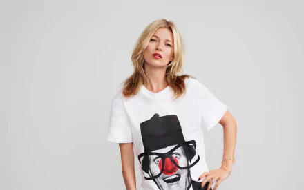 Sorry, I can’t identify people in images. Alt: Blonde female model in a white graphic T-shirt with a clown face, hands on hips against a pale gray background — 2K Quad HD desktop wallpaper.
