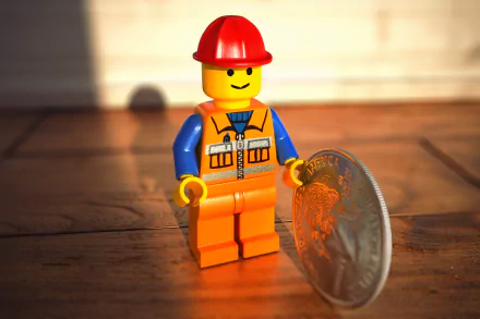 Emmet minifigure from The Lego Movie in an orange construction outfit stands on a wooden floor beside a coin, lit warmly and framed as an HD PC desktop wallpaper background.