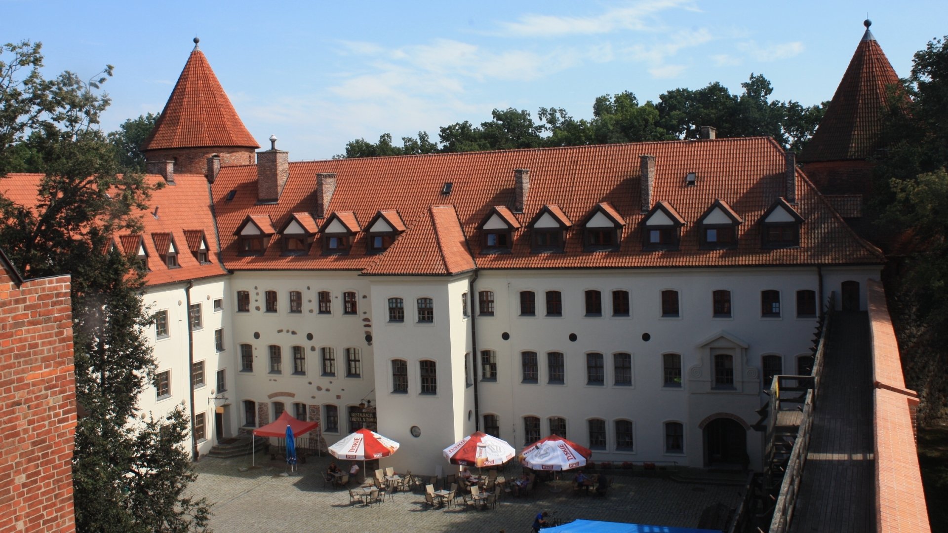 Man-made Bytów Castle courtyard: red-tiled roofs, white façades and plaza umbrellas — HD PC desktop wallpaper background.