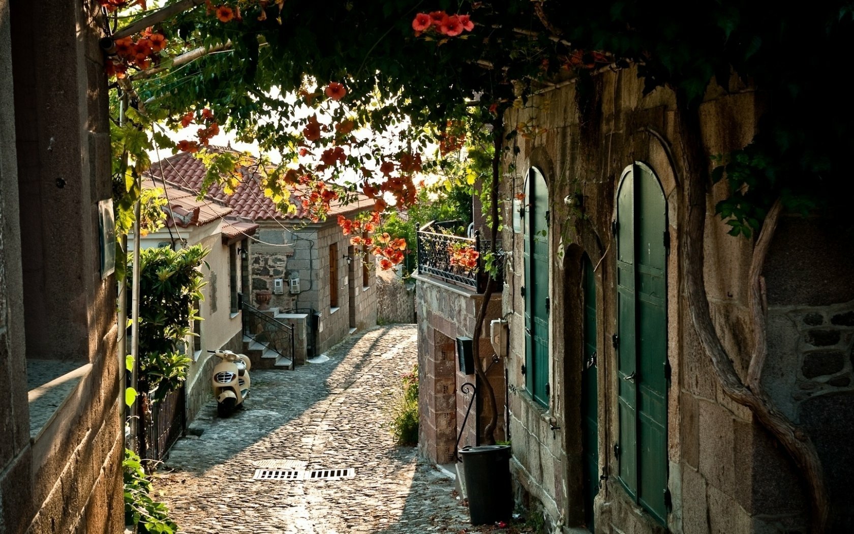 HD PC desktop wallpaper showing a charming man-made alley with cobblestone path, stone buildings, and vibrant hanging flowers, bathed in warm sunlight.