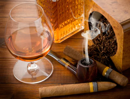 HD desktop wallpaper featuring a glass of brandy, a lit smoking pipe, cigars, and a box of tobacco on a wooden table. The rich brown tones and cozy setup create a warm, inviting atmosphere.