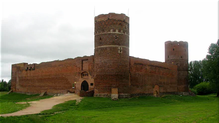HD desktop wallpaper showing the man-made red brick Ciechanów Castle with round towers and surrounding green grass under a cloudy sky.