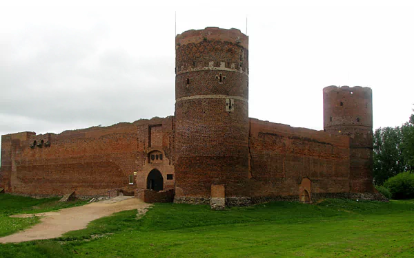 HD desktop wallpaper showing the man-made red brick Ciechanów Castle with round towers and surrounding green grass under a cloudy sky.