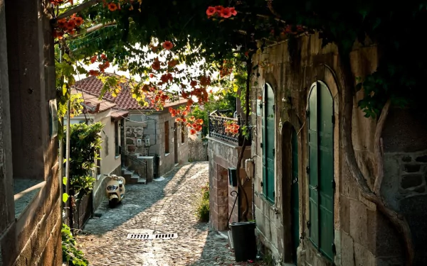 HD PC desktop wallpaper showing a charming man-made alley with cobblestone path, stone buildings, and vibrant hanging flowers, bathed in warm sunlight.