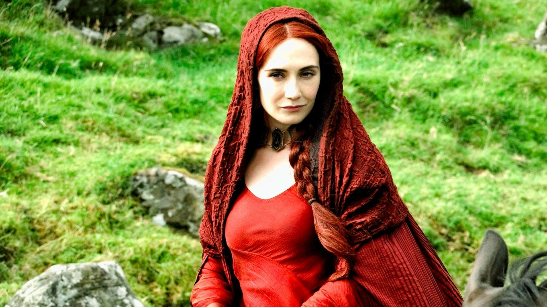 A striking HD wallpaper featuring Melisandre from Game of Thrones, portrayed by Carice van Houten, captures her in a vivid red gown against a lush green backdrop.