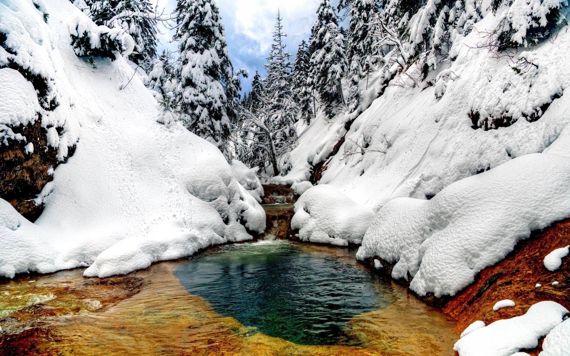 A tranquil winter scene featuring a snowy landscape with a clear stream flowing between snow-covered banks and evergreen trees, set against majestic mountains.