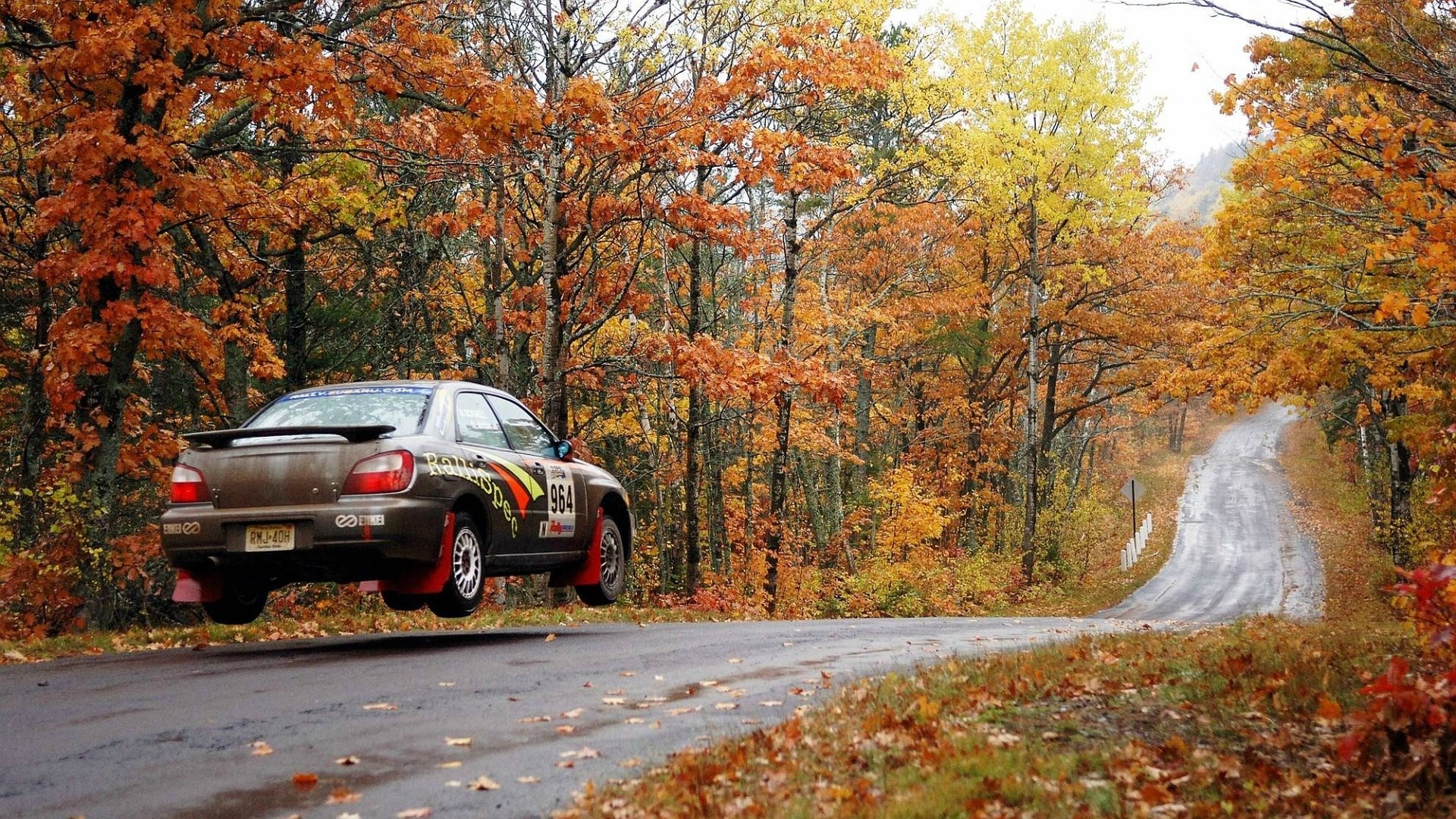 HD PC desktop wallpaper and background showing a Subaru Impreza rally car airborne on a winding country road through a vibrant autumn forest.