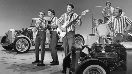 Black-and-white HD PC desktop wallpaper background showing a 1960s surf‑rock band performing on a studio set with guitars, drums and vintage hot rods — music-themed vintage scene.