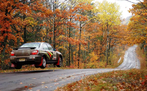 HD PC desktop wallpaper and background showing a Subaru Impreza rally car airborne on a winding country road through a vibrant autumn forest.
