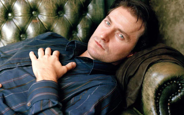 HD desktop wallpaper featuring English actor Richard Armitage reclining on a vintage leather sofa, dressed in a dark striped shirt with a relaxed expression.