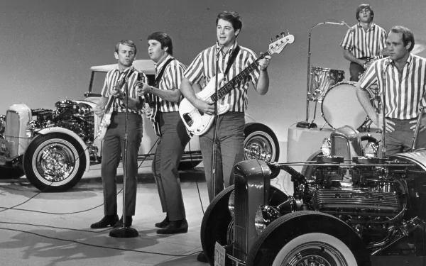 Black-and-white HD PC desktop wallpaper background showing a 1960s surf‑rock band performing on a studio set with guitars, drums and vintage hot rods — music-themed vintage scene.