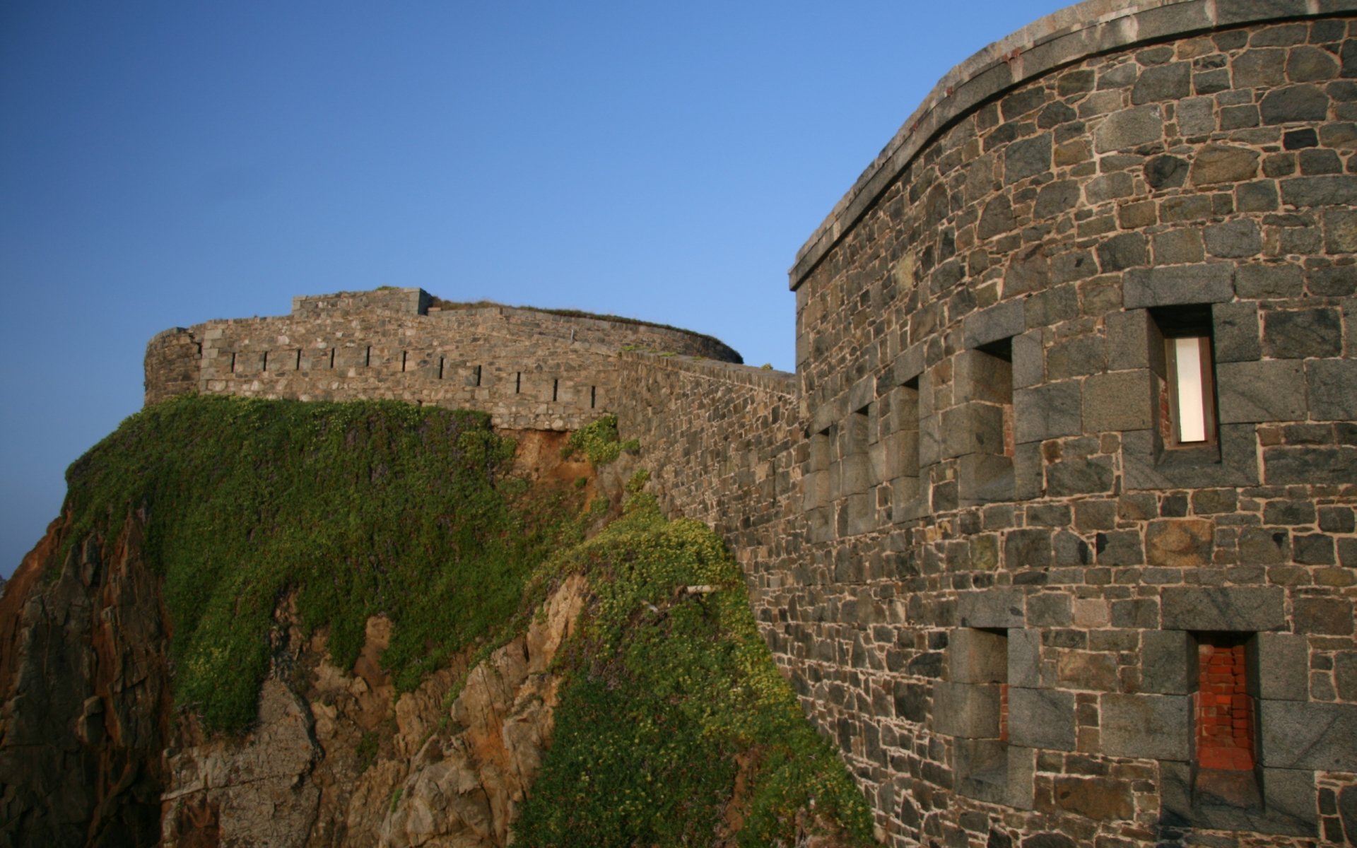 A stunning HD desktop wallpaper showcasing Fort Clonque, featuring its impressive stone architecture against a clear blue sky, nestled atop a lush green cliff.