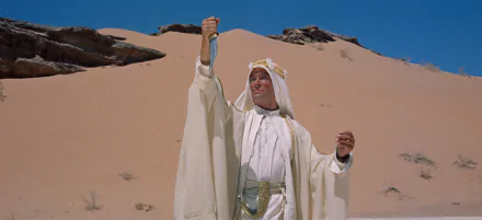 HD PC desktop wallpaper: Lawrence of Arabia movie scene of a robed man raising a dagger against vast sand dunes under a clear blue sky.