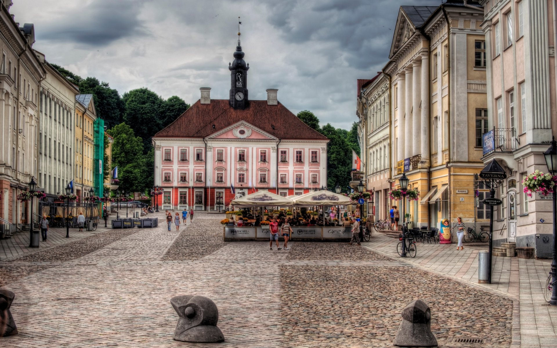2K Quad HD PC desktop wallpaper of Tartu’s cobblestone town square, showcasing man-made historic architecture: the pink town hall, flanked by classical buildings, market stalls and an overcast sky.