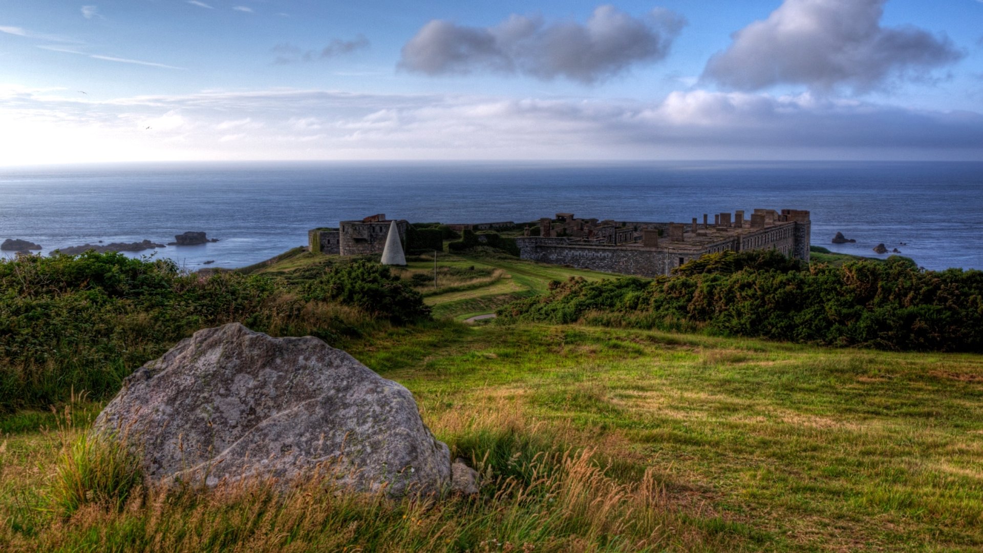 HD desktop wallpaper of Fort Tourgis, a man-made coastal fortification surrounded by grassy fields under a cloudy sky.