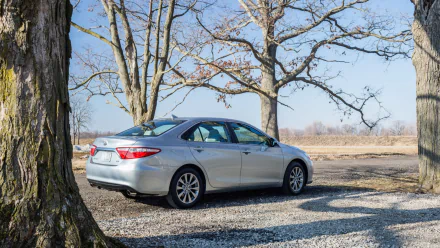 HD PC desktop wallpaper: silver 2015 Toyota Camry parked on a gravel drive beneath leafless trees in a rural landscape under a clear blue sky.