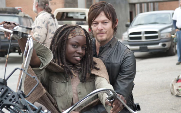 Image from the TV show The Walking Dead featuring two characters posed on a motorcycle, set against a backdrop of parked vehicles in a gritty urban environment.