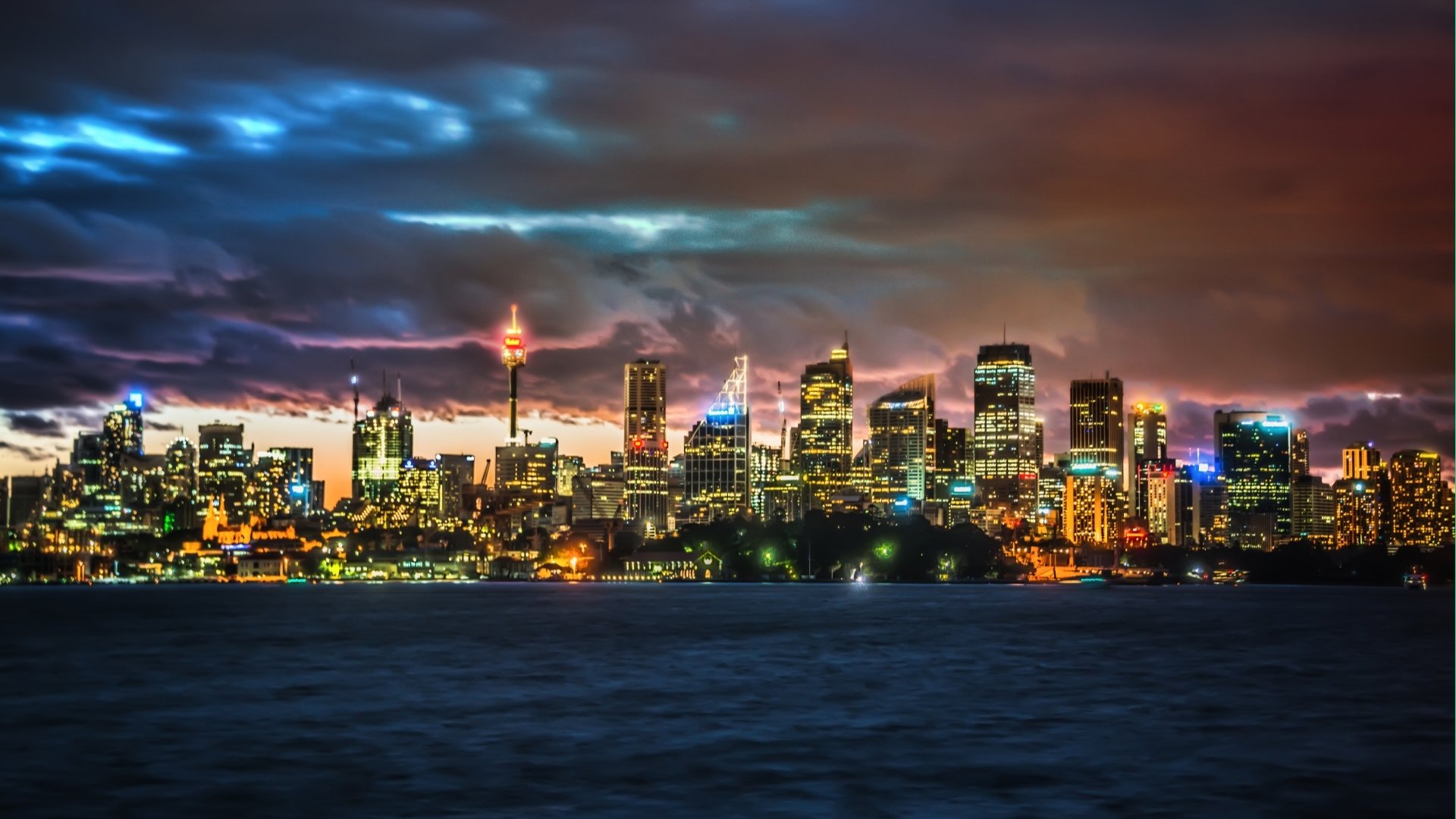 HD PC desktop wallpaper: vibrant nighttime Sydney skyline with illuminated skyscrapers, cloudy sky and glowing city lights reflecting on the harbor — man-made urban panorama.