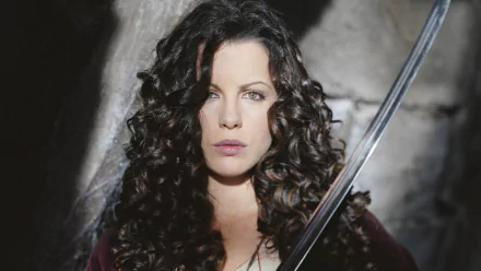 Anna Valerious, portrayed by Kate Beckinsale in the movie Van Helsing, gazes intently while holding a sword, set against a dramatic background. HD desktop wallpaper.