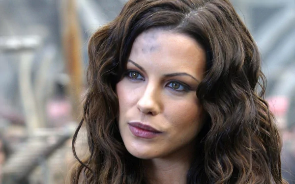 A close-up of a woman with wavy brown hair and striking blue eyes, featuring a backdrop from the movie Van Helsing, designed as a HD PC desktop wallpaper.