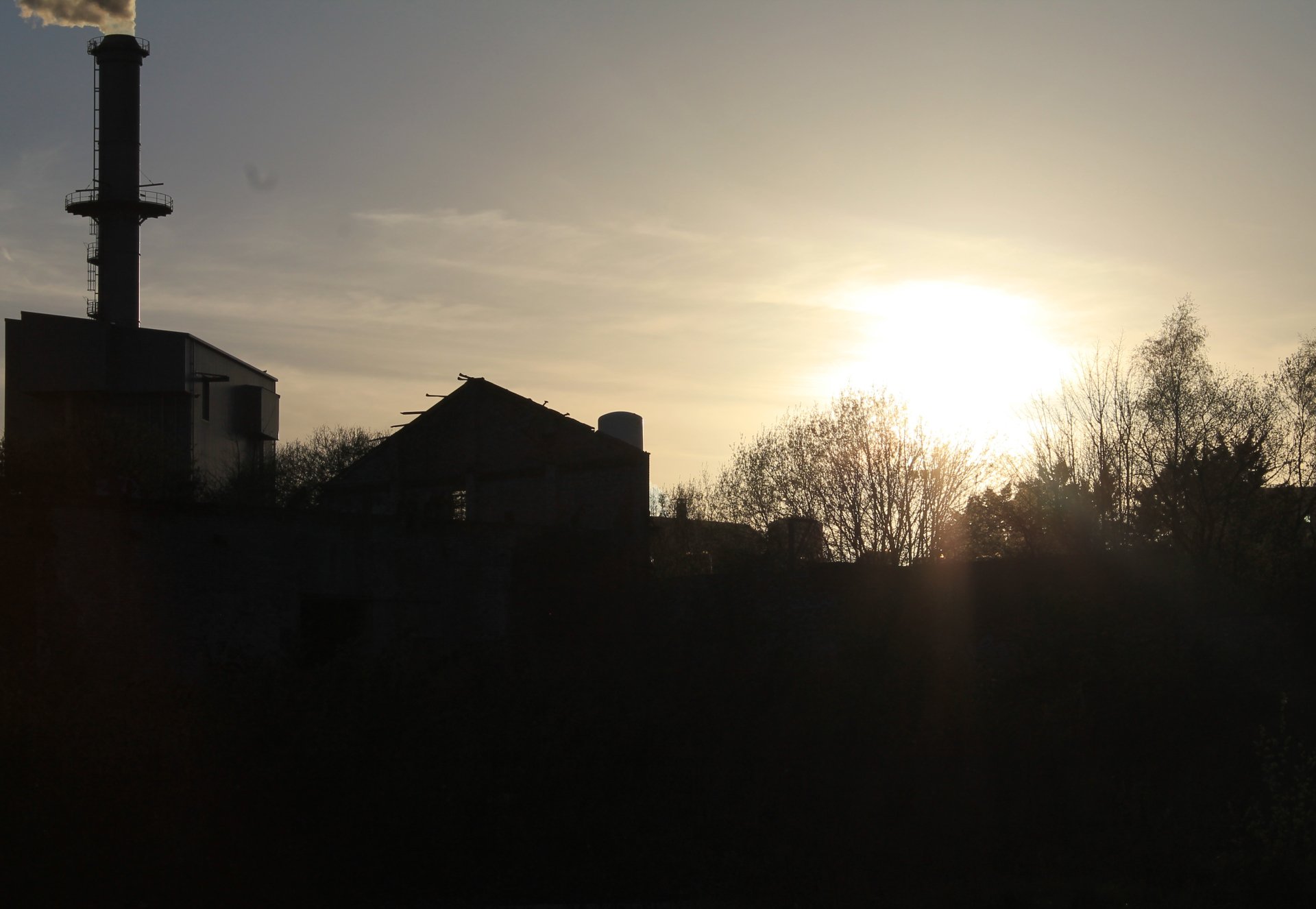 4K Ultra HD PC desktop wallpaper/background: industrial skyline and man-made buildings silhouetted at sunset, smokestack and treeline beneath a glowing low sun.