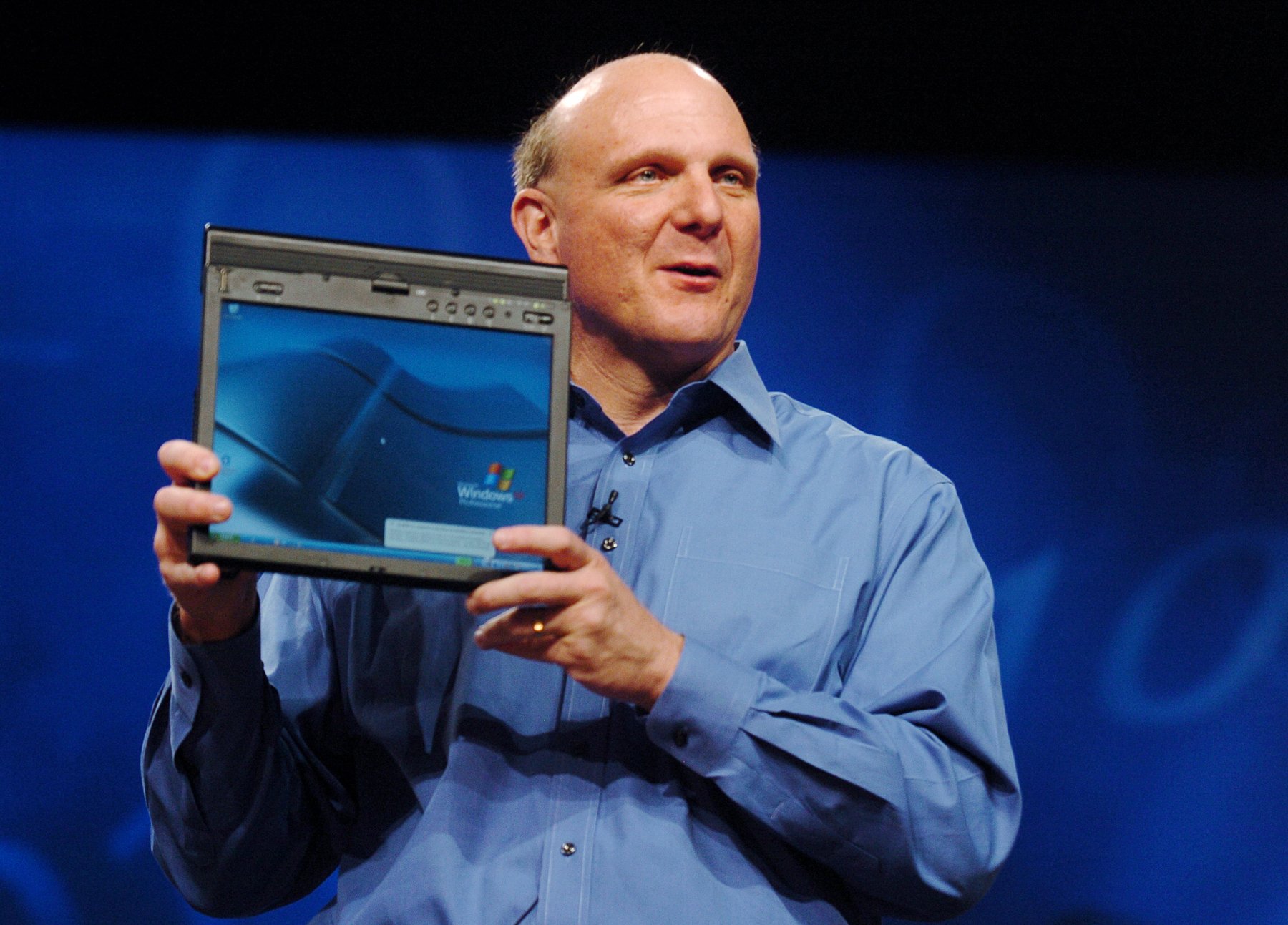 HD desktop wallpaper showing a man in a blue shirt holding up a software package on a stage with a deep blue background.