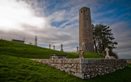 Clonmacnoise Monastery round tower and stone crosses over a grassy cemetery in Ireland under a dramatic sky — HD desktop wallpaper.