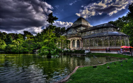 HD PC desktop wallpaper: man-made Palacio de Cristal glass pavilion beside a reflective pond, lush greenery and dramatic sky.
