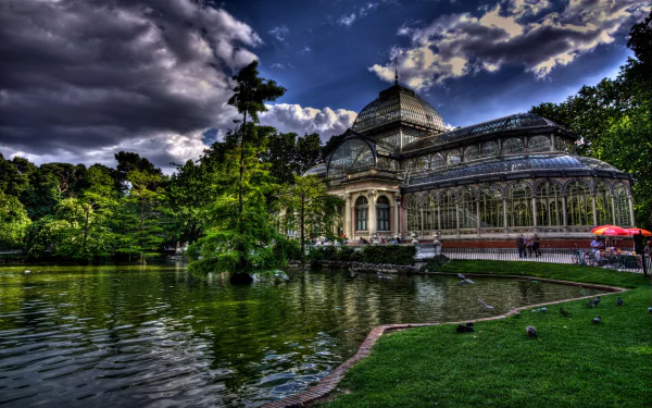 HD PC desktop wallpaper: man-made Palacio de Cristal glass pavilion beside a reflective pond, lush greenery and dramatic sky.