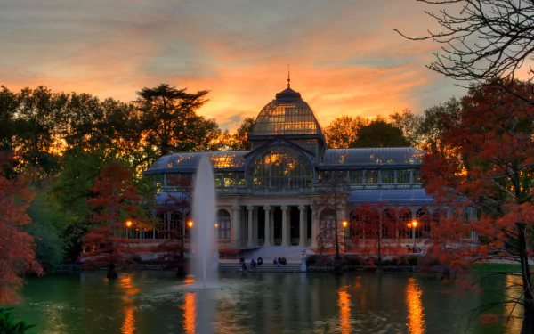 HD desktop wallpaper showcasing the Palacio de Cristal, a man-made glass structure, illuminated at sunset with vibrant autumn trees and a water fountain in the foreground.