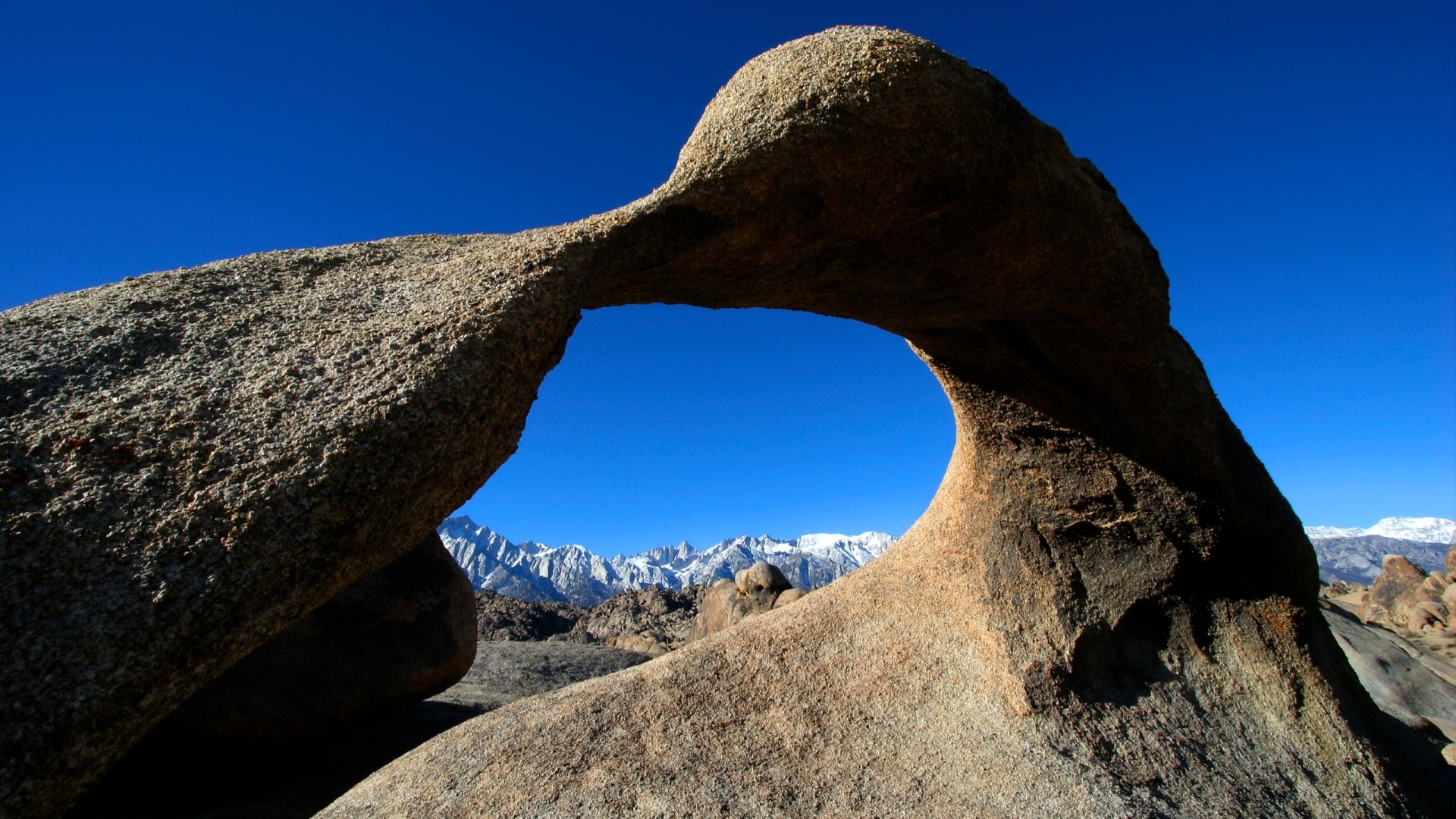 Mobius Arch nature scene: rocky natural arch framing snow-capped peaks under a deep blue sky — 2K Quad HD PC desktop wallpaper and background.