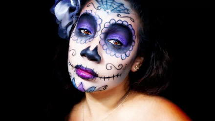 Artistic sugar skull portrait with purple eye makeup and floral hair accent on a black background — 2K Quad HD PC desktop wallpaper and background.