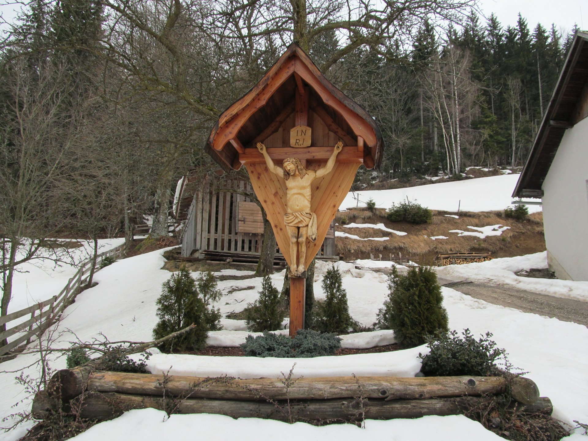 A wooden cross with a carved figure of Jesus is displayed under a shelter in a snowy landscape, surrounded by greenery and rustic buildings, serving as a serene religious focal point.