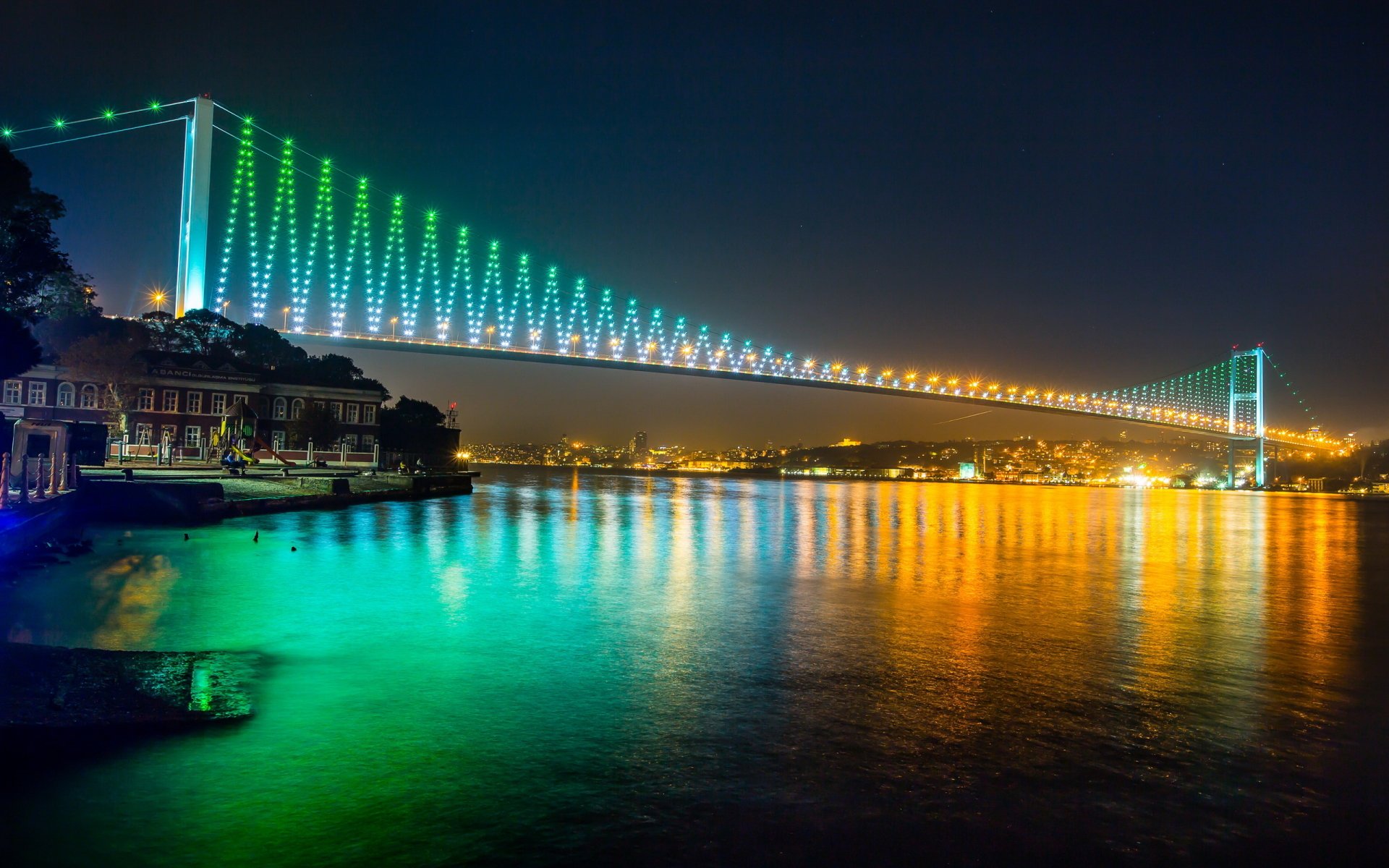 HD desktop wallpaper of the Bosphorus Bridge illuminated at night, showcasing vibrant green lights reflecting on the water beneath this iconic man-made structure.