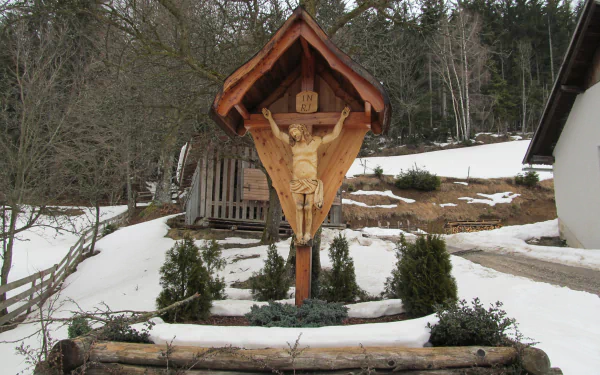 A wooden cross with a carved figure of Jesus is displayed under a shelter in a snowy landscape, surrounded by greenery and rustic buildings, serving as a serene religious focal point.