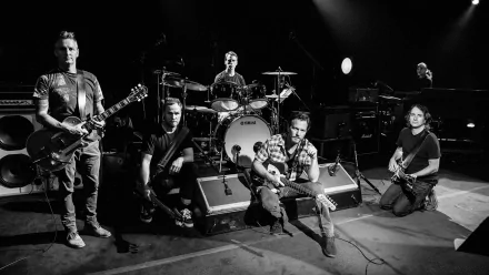 Black-and-white HD PC desktop wallpaper of a rock music band onstage: five members with guitars and drums, some seated, some standing under spotlights, captured mid-performance.