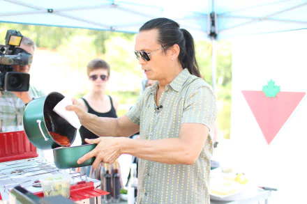 Chef at an outdoor event pouring sauce, with a camera crew in the background, in a HD desktop wallpaper format.