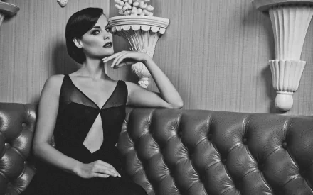 Black and white HD wallpaper of American actress Jaimie Alexander elegantly posed on a tufted leather sofa with ornate wall decor in the background.